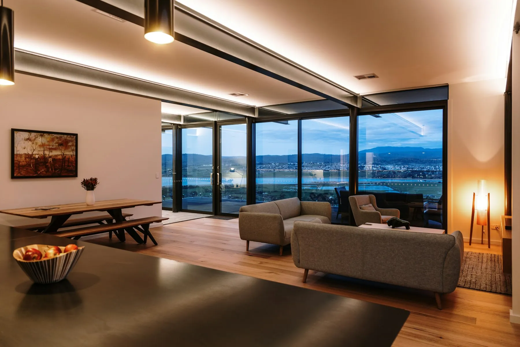 The open-plan living and dining space, with a long timber table and a wall of glass over the water